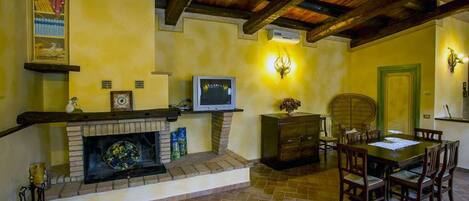 A cozy room with a fireplace, wooden ceiling beams, a dining table with chairs, and a television on a shelf.