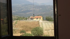 Blick aus dem Fenster auf eine ländliche Landschaft mit einem gelben Haus, einem Feld und fernen Bergen.