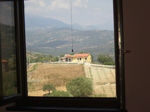 A window view of a rural landscape with a yellow house, a dirt field, and distant mountains.