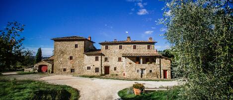 Un bâtiment en pierre doté de nombreuses fenêtres et d’une porte rouge, entouré de verdure et bordé d’un chemin de gravier.