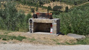 Un área de estar al aire libre rústica con un banco de piedra y una mesa de metal, rodeada de olivos y un paisaje montañoso.