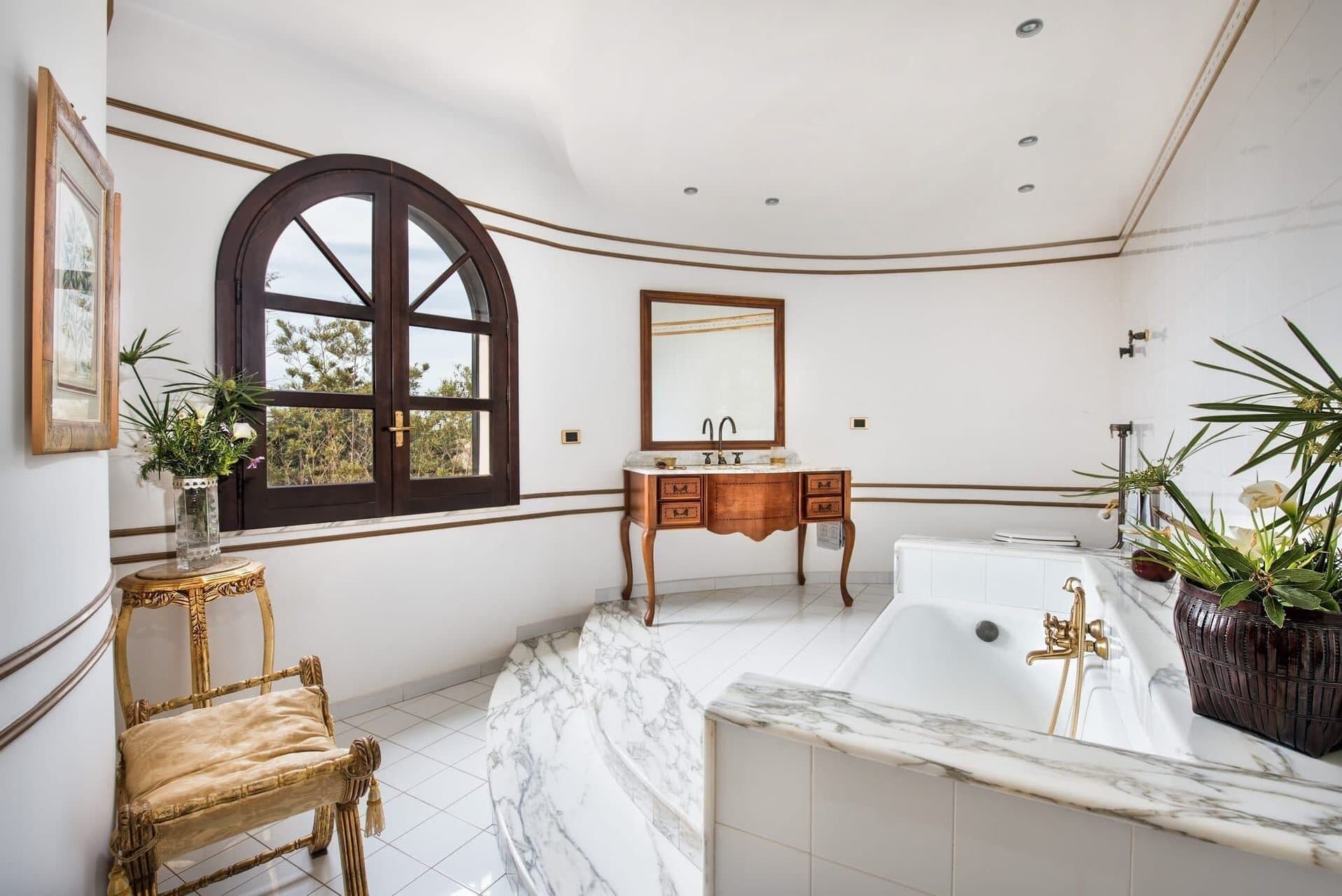 A spacious bathroom with a large arched window, a wooden vanity, a marble bathtub, and a wicker chair.