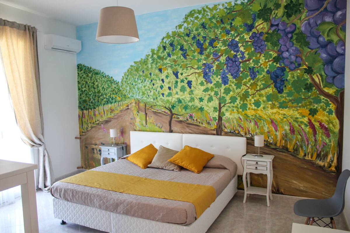 Un dormitorio con una cama grande, dos mesitas de noche, una silla y un mural en la pared de un viñedo.