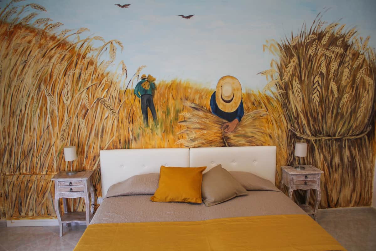 Un dormitorio con una cama grande, dos mesitas de noche y un mural que representa un campo de trigo con dos figuras.