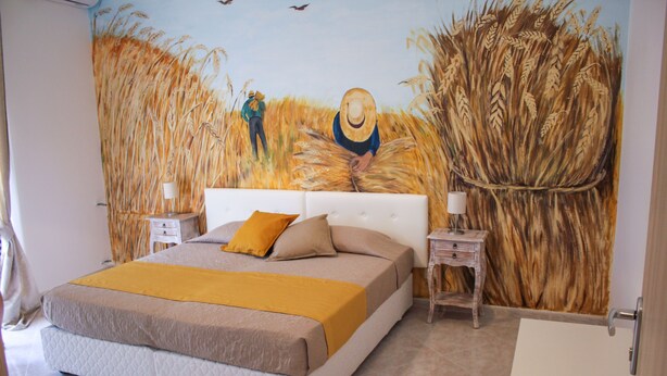 A bedroom with a large bed, two bedside tables, a nightstand, and a wall mural of a wheat field. - Belice Rooms (Salemi)
