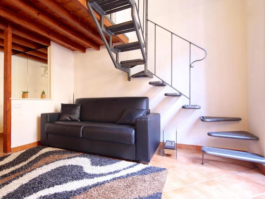 Ein modernes Wohnzimmer mit einer schwarzen Ledersofa, einem gemusterten Teppich, einer Treppe mit Metallgeländer und einem kleinen Tisch.