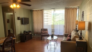 Una sala de estar con comedor, ventilador de techo, televisor, sofá, mesa de centro y vistas al exterior.