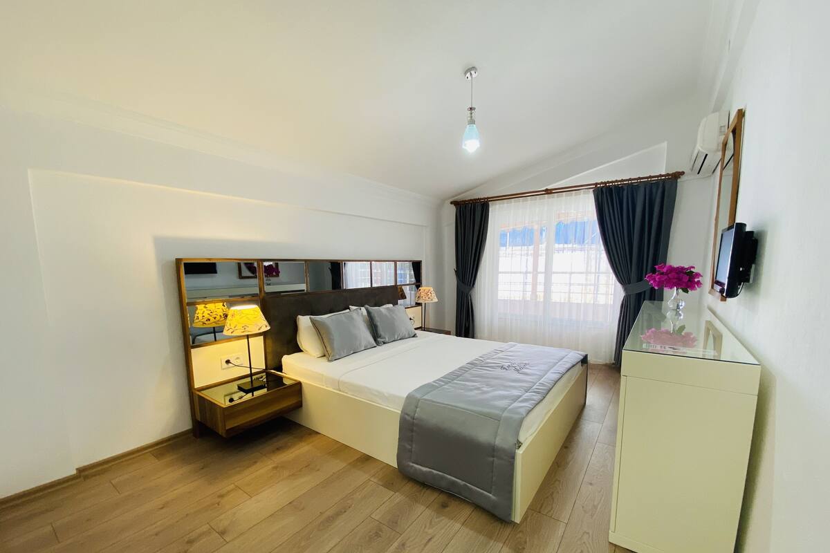 A bedroom with a bed, bedside tables, a nightstand, a dresser, a mirror, and a window with curtains.