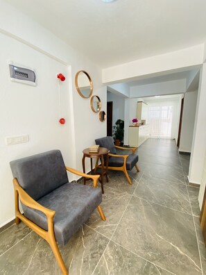 A hotel lobby with a grey sofa, wooden chairs, a small round table with a book, and a potted plant.