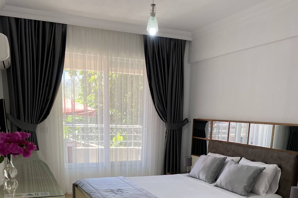 A hotel room with two beds, a bedside table, a lamp, a window with curtains, and a view of greenery outside.