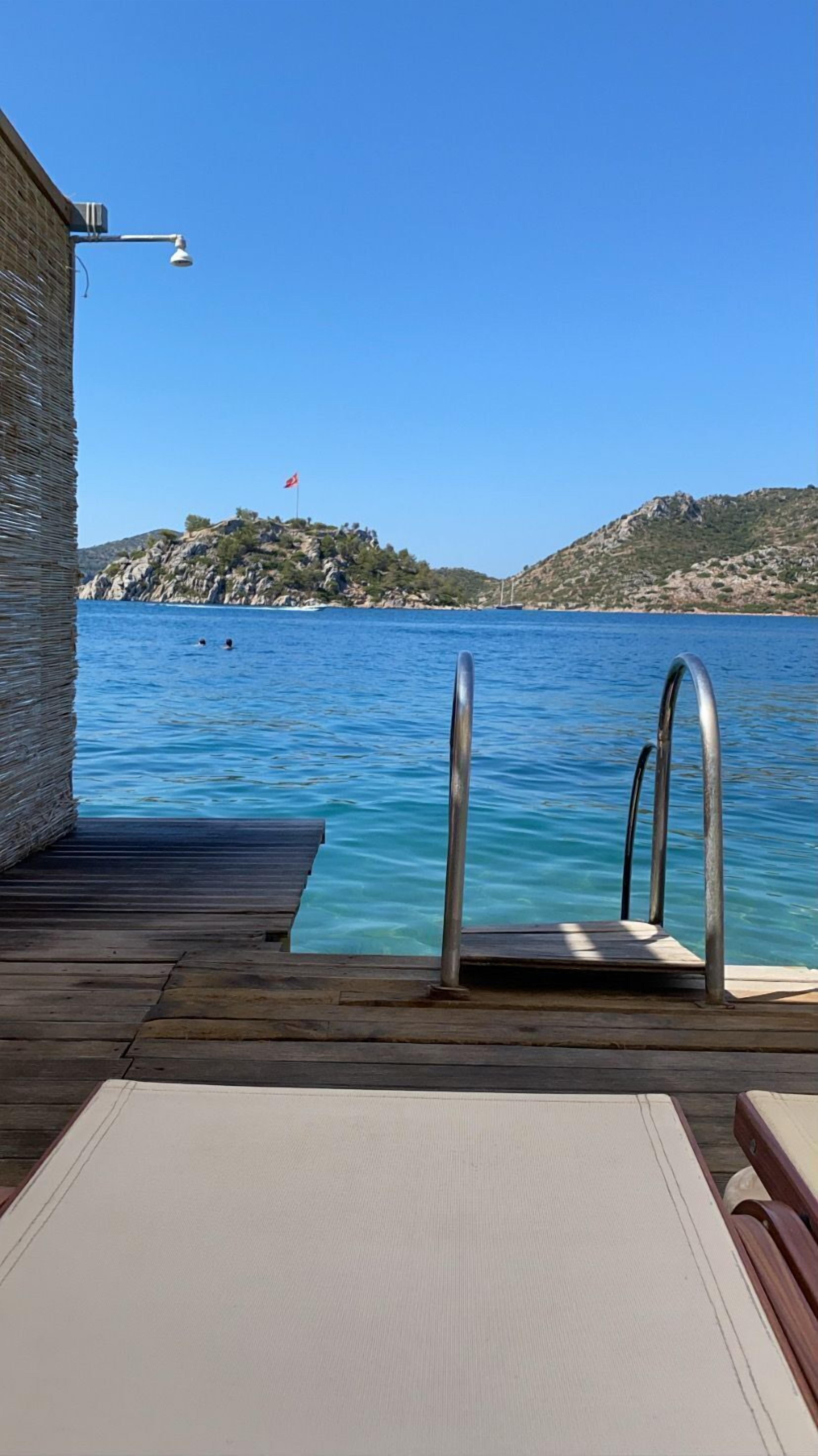 Una terraza de madera que da a una piscina con barandillas de metal, con vistas al mar azul y a colinas rocosas.