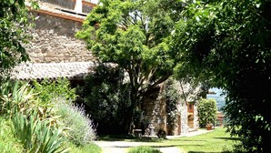 Un bâtiment en pierre, surmonté d’un toit de tuiles, entouré d’une végétation luxuriante et accessible par un chemin dégagé menant à un banc.