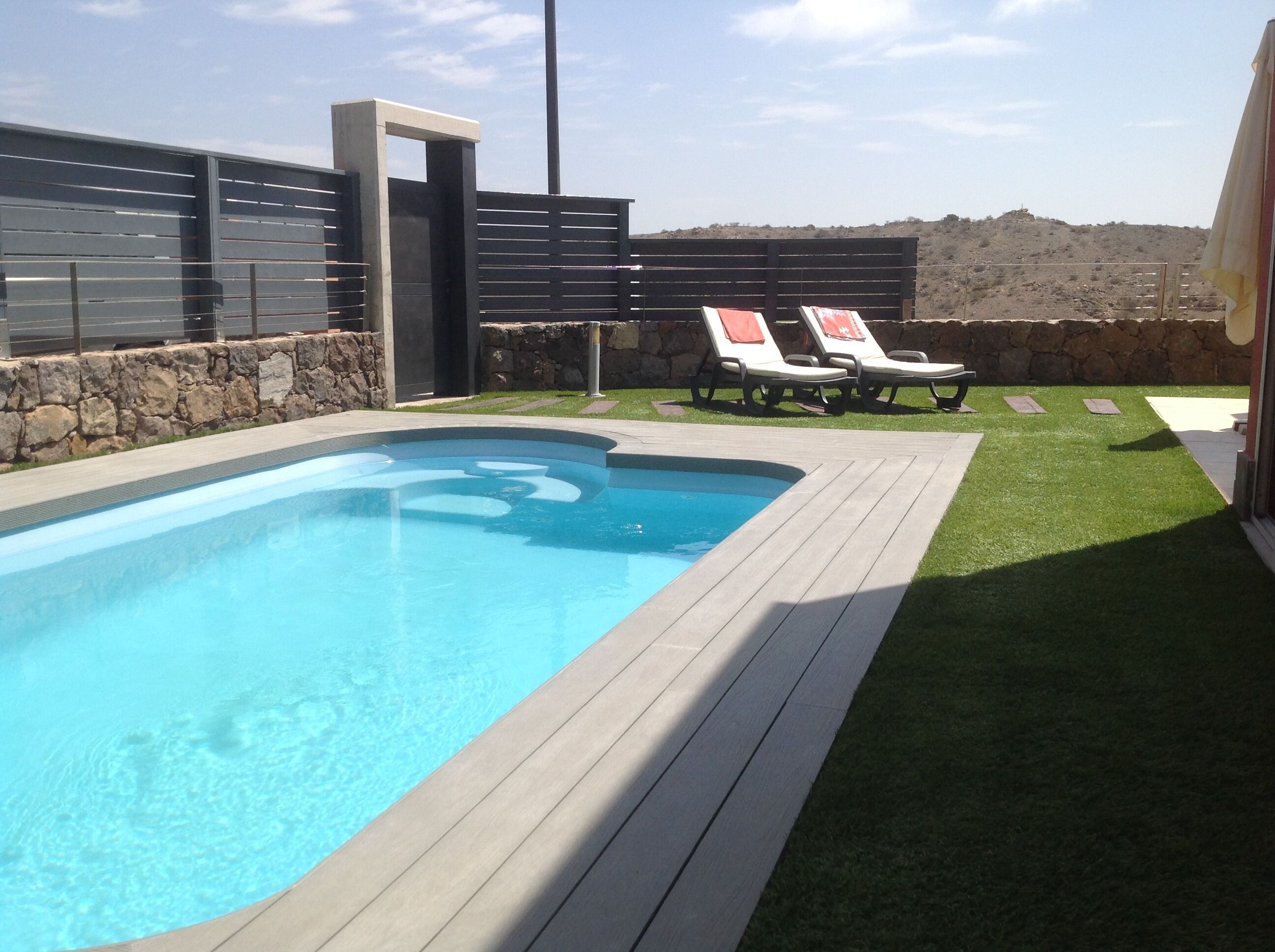 A swimming pool with a wooden deck, two lounge chairs, and a stone wall.