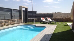 Una piscina con cubierta de madera, dos sillas reclinables y un muro de piedra.