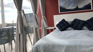 Una habitación de hotel con cama, un ventanal con cortinas, vistas al cielo y un cuadro en la pared.