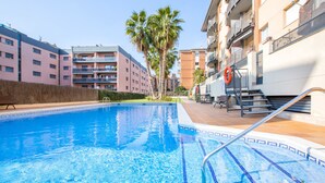 A swimming pool with clear blue water, surrounded by a tiled deck. There are palm trees and a building with balconies in the background. - 2P53 - Cozy apartment for 4 people with pool located near the center and the beach of Fenals (Lloret de Mar) (Lloret de Mar)
