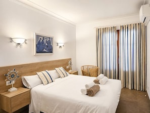 A hotel room with a large bed, two bedside tables with decorative items, a wall-mounted light fixture, a framed artwork, and a window with striped curtains.