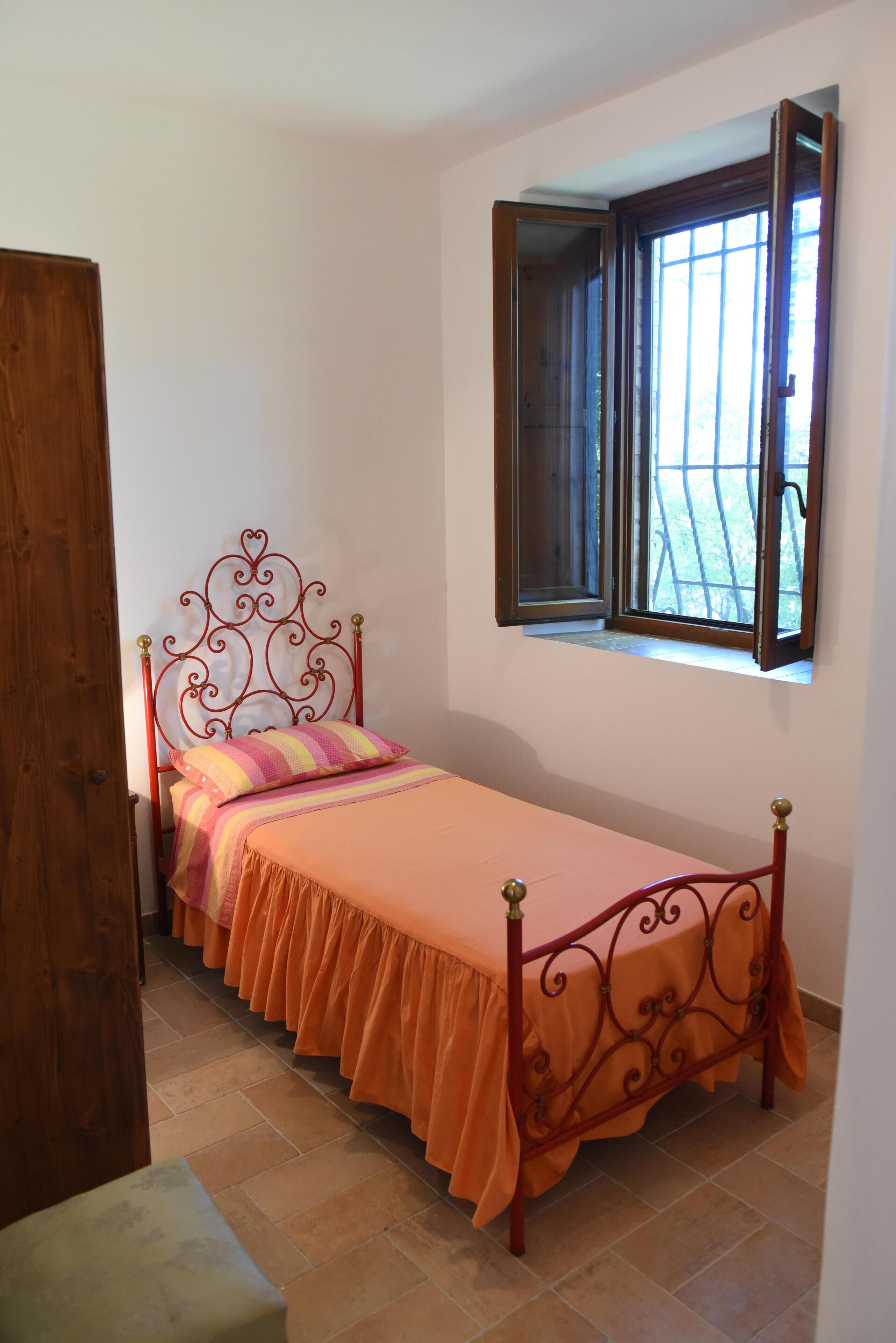 Un lit simple avec une couvre-lit orange et un oreiller à rayures, dans une pièce dotée d’une fenêtre et d’une porte en bois.