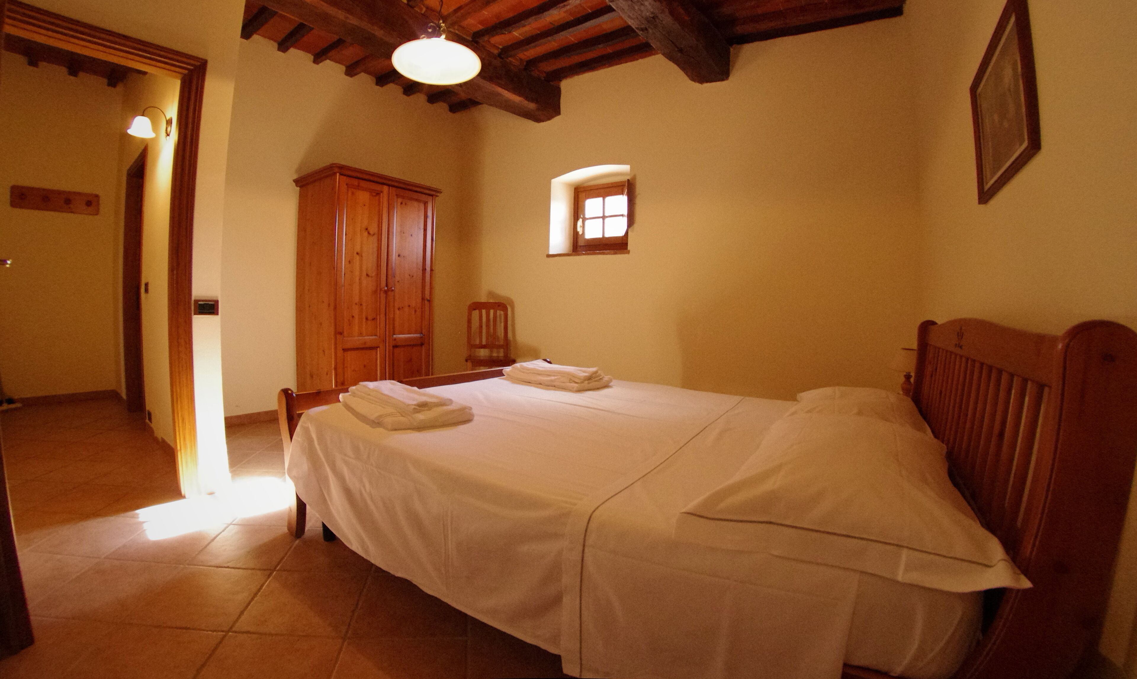 A bedroom with a bed, a wooden wardrobe, a small window, and a door leading to another room.