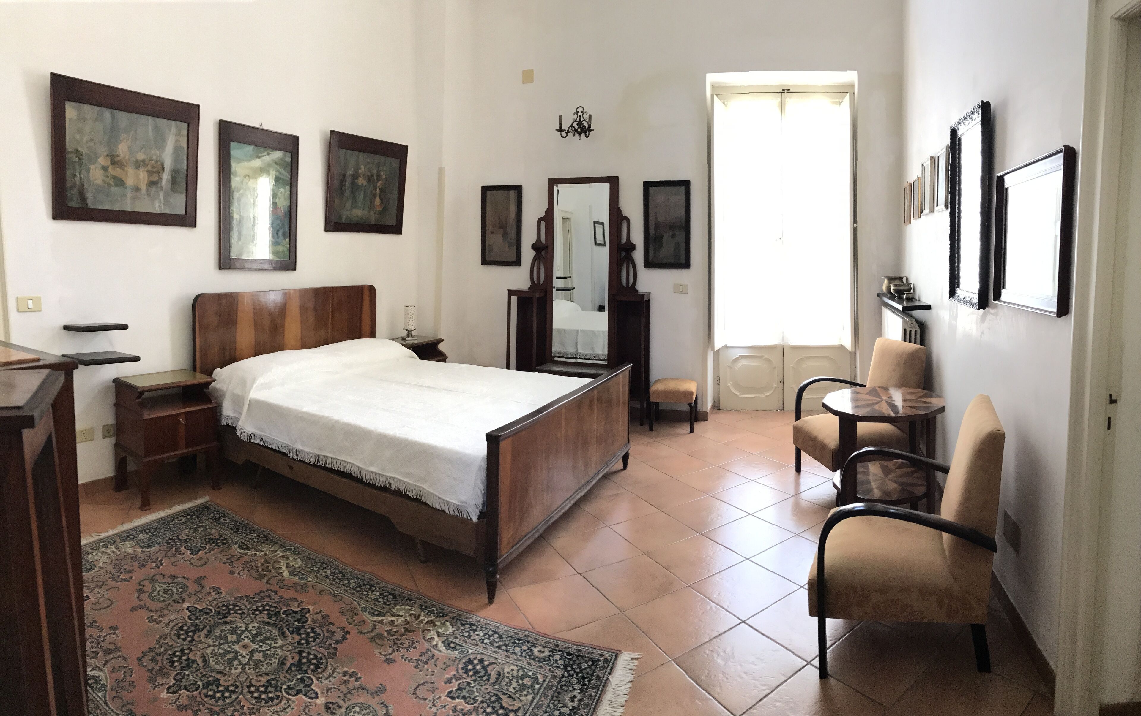 A bedroom with a wooden bed, a small bedside table, a chair, a round table, a mirror, and three framed pictures on the wall.