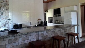 A modern kitchen with white cabinets, a granite countertop, and a wooden ceiling. There are two wooden stools and a coffee machine on the counter. - São Roque House in Paraty (São Roque)