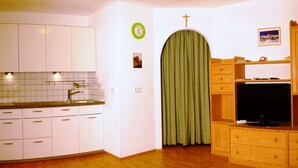 Une cuisine de taille réduite avec des meubles blancs, un rideau vert, une horloge et un téléviseur posé sur un meuble en bois.