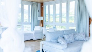 A modern living room with a sofa, a chair, and a lamp. There are large windows with curtains. - Solano Penthouse – Los Cuatro Vientos Rural Retreat (Moratalla)