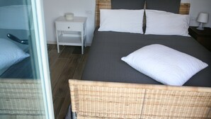 Ein ordentlich bezogenes Bett mit grauer Tagesdecke und weißen Kissen, ein kleiner Nachttisch mit Lampe sowie ein Korbstuhl.