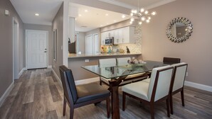 A dining area with a glass-top table, four chairs, a chandelier, and a kitchen in the background. - Beautiful 1st. floor unit totally renovated with lake view! (Naples)
