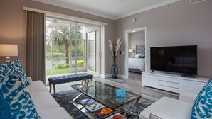 A modern living room with a glass coffee table, a flat-screen TV, and a large window offering a view of greenery. - Beautiful 1st. floor unit totally renovated with lake view! (Naples)