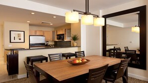 A modern dining area with a wooden table, chairs, and a fruit bowl. There is a kitchen in the background with wooden cabinets and a refrigerator.