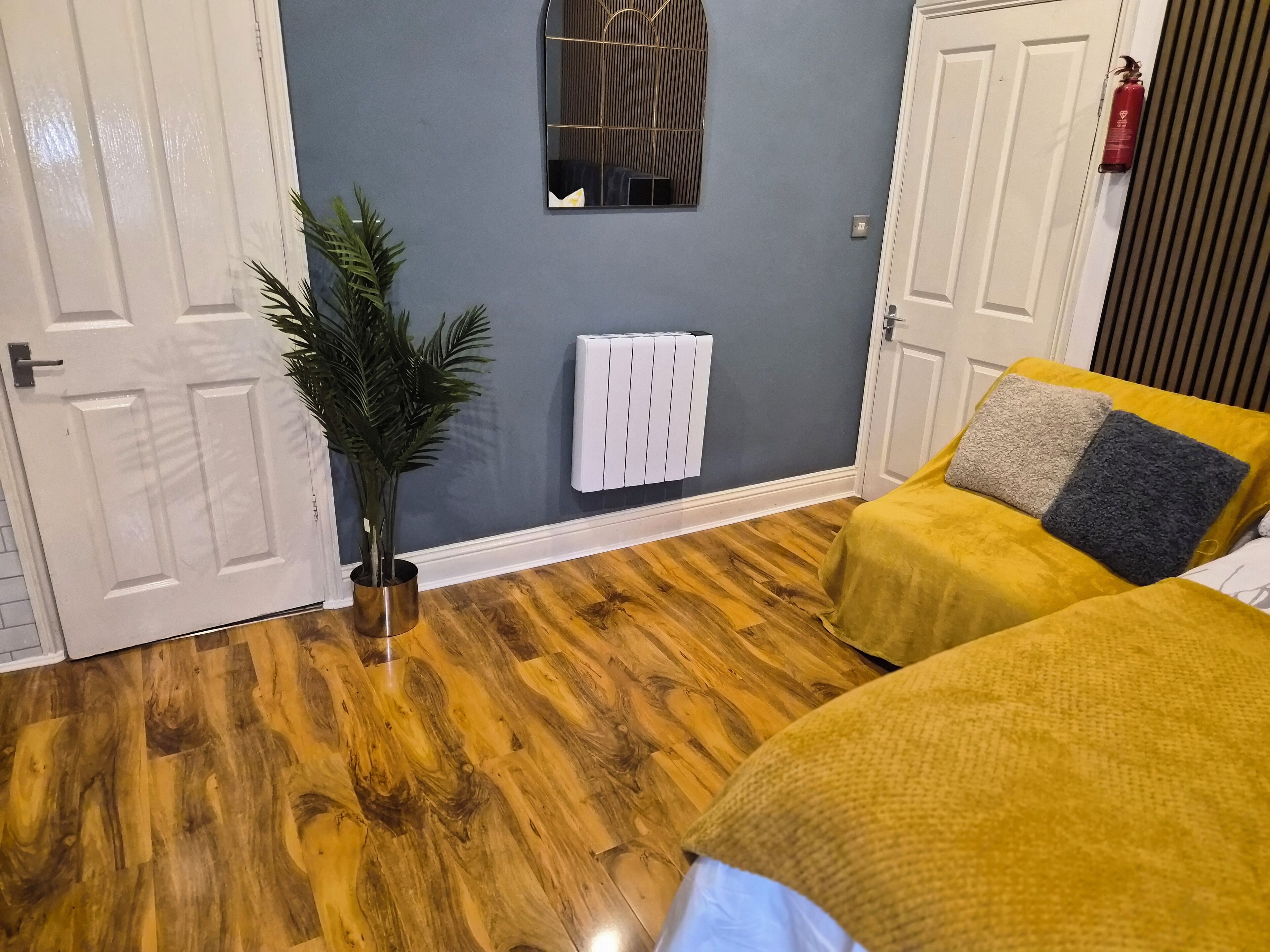 A room with a wooden floor, a radiator, a potted plant, and a bed with a yellow bedspread and two pillows.