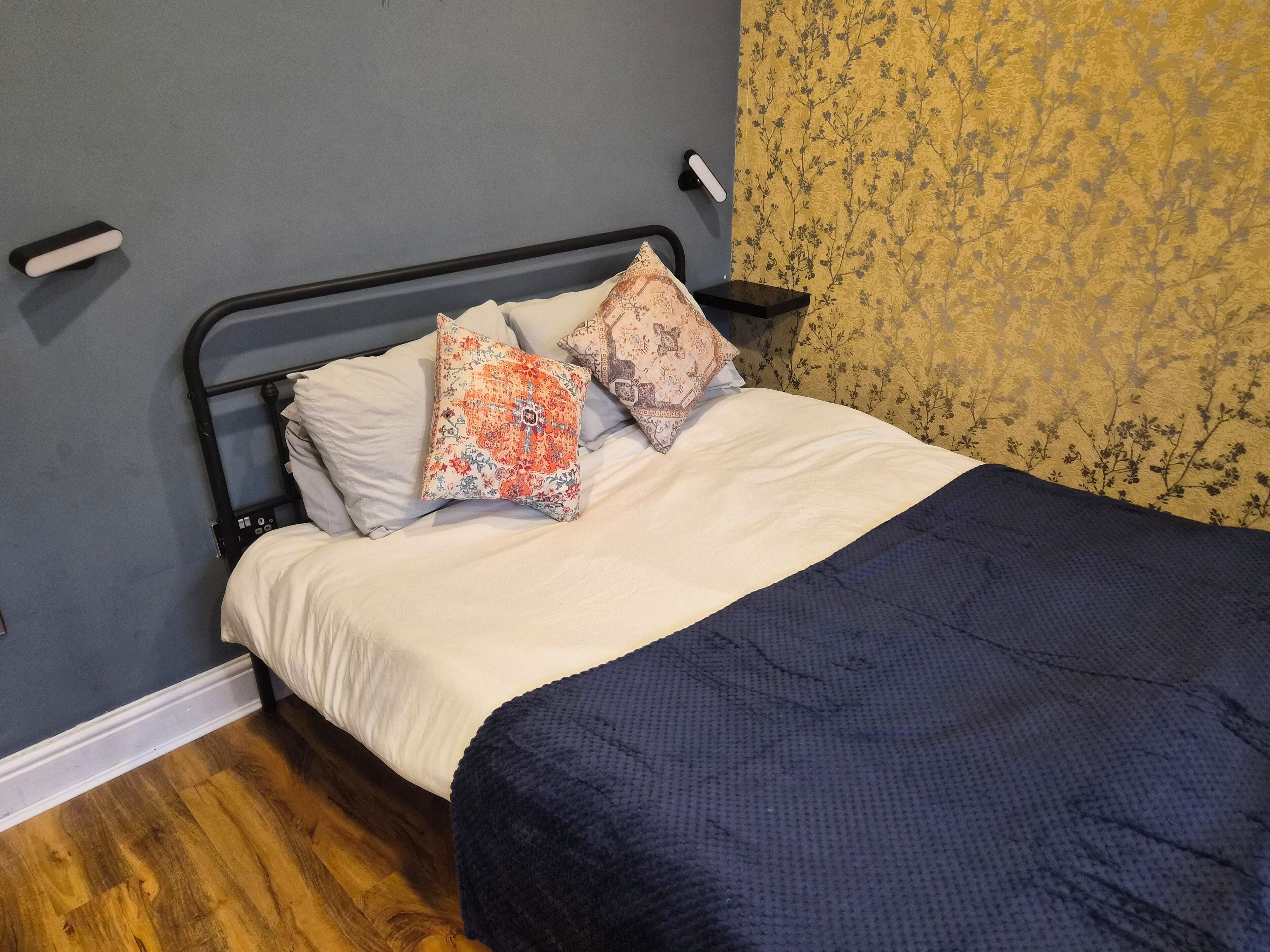 Una cama con un edredón azul, dos almohadas estampadas y una almohada blanca, sobre un suelo de madera.