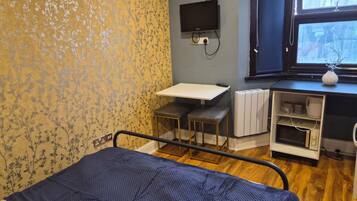 Ett sjukhusrum med en säng, ett litet bord, en stol, en tv som är monterad på väggen, ett fönster med utsikt över träd och ett element.