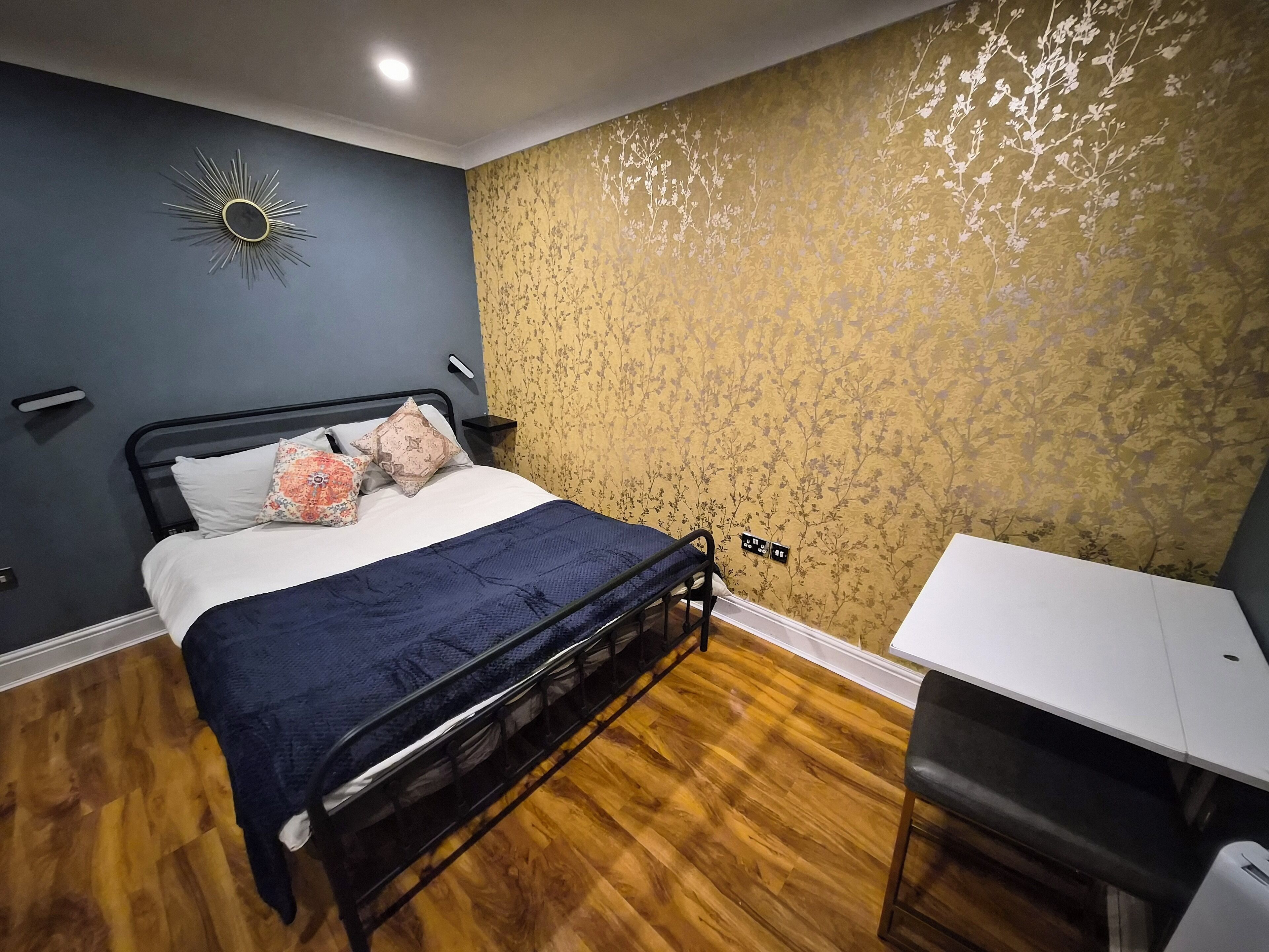 A bedroom with a bed, a desk, a chair, a nightstand, a wall clock, and a power outlet.