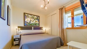 A bedroom with a bed, bedside tables, a nightstand, a painting, a window with curtains, and a view of buildings outside. - AMBAR 18B - Apartment with sea views very close to the beach and supermarkets (Calpe)