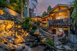 Eine steinerne Stützmauer mit Wasserspiel, eine Holzterrasse und ein Gebäude mit Balkon und Außenbestuhlung.