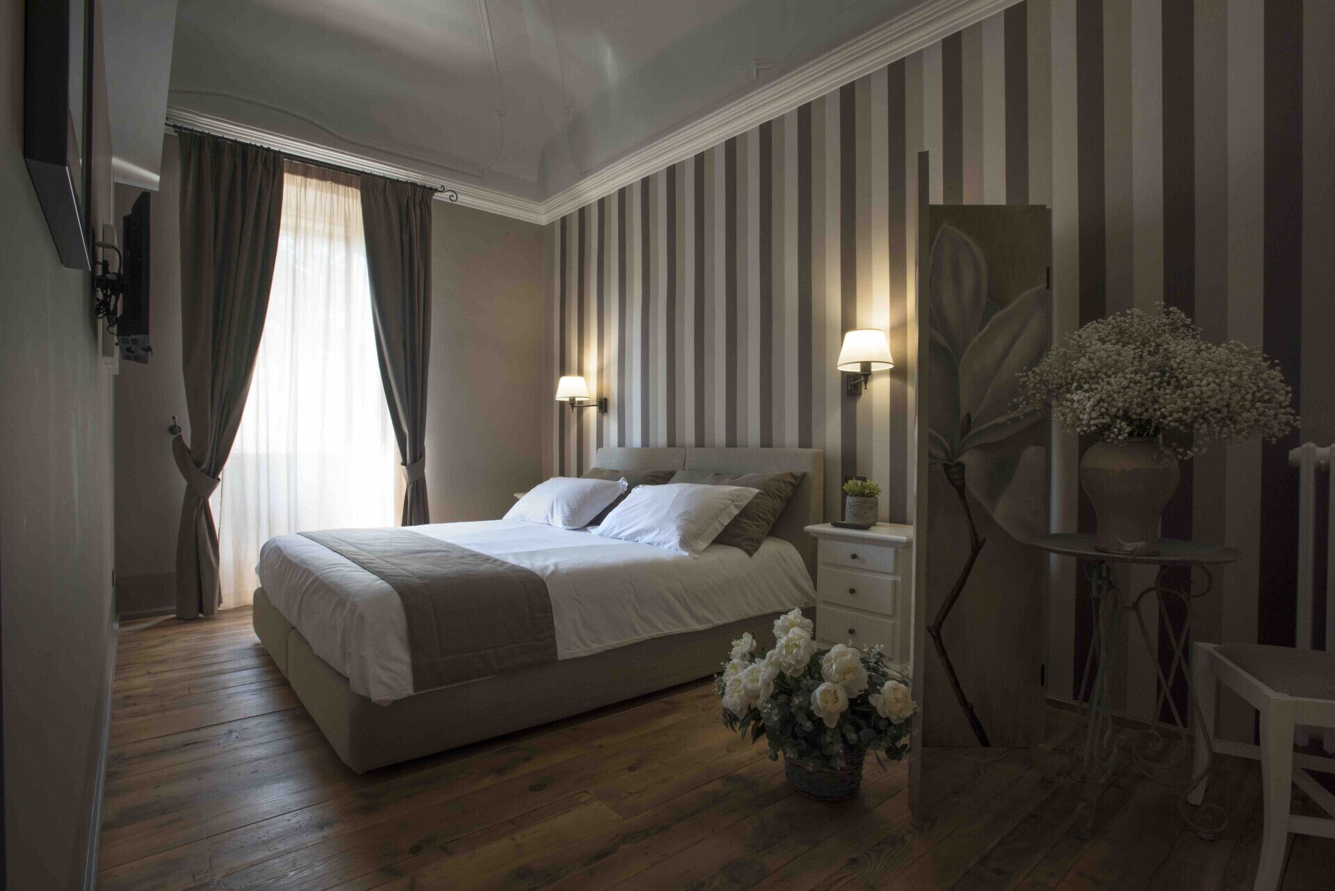 A bedroom with a large bed, two bedside lamps, a nightstand, a chair, a potted plant, and a striped wall.