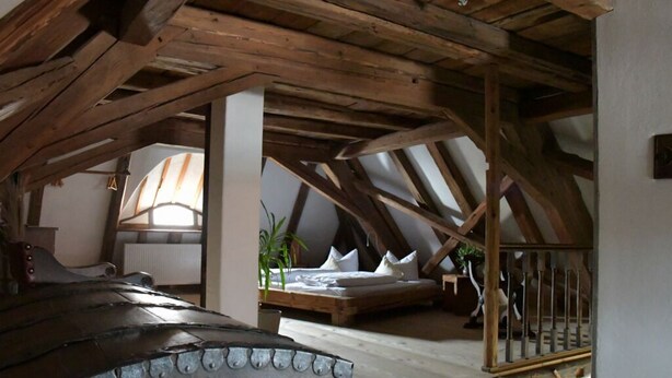 Ein Schlafzimmer mit schrägen Wänden, Holzbalken, einem Bett und einem kleinen Balkon.