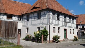 Ein traditionelles Fachwerkhäuschen mit einem steil geneigten roten Ziegeldach, einer grünen Tür und einem kleinen Garten mit blühenden Pflanzen.