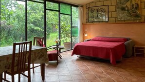 Ein Zimmer mit Bett, Tisch, Stühlen und Blick auf Grün hinter großen Fenstern.