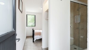Ein Badezimmer mit Glasdusche, einem Bett mit weißer und beiger Decke und einem Fenster mit Grünpflanzen vor der Ansicht.