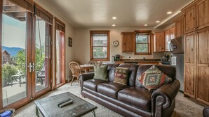 A spacious living area with a leather sofa, dining table, and chairs. A sliding glass door leads to a balcony with a view of a mountain and trees. The kitchen features wooden cabinets and a refrigerator. - Ground-Floor Unit w/Views, Close to Trails & Golf! (Keystone)