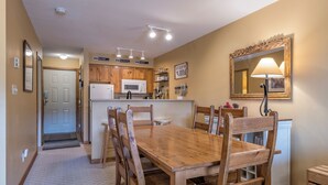 A dining area with a wooden table and chairs, a kitchen with wooden cabinets, a microwave, and a sink. There is a mirror on the wall and a lamp on the table.