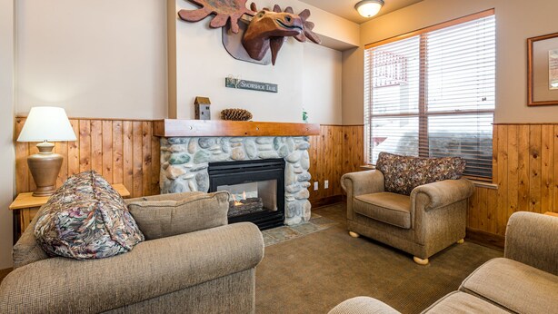 A cozy living room with a stone fireplace, wooden walls, and a mounted deer head sculpture. There are two armchairs and a lamp on a side table.