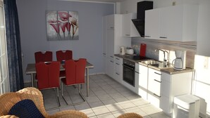 A modern kitchen with white cabinets, a built-in oven, and a coffee maker. There is a dining area with red chairs and a table. - Holiday apartments in the middle of the biosphere reserve (Middelhagen)