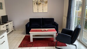 A living room with a dark blue sofa, a white coffee table, a red rug, a black chair, and a TV on a white cabinet. - Holiday apartments in the middle of the biosphere reserve (Middelhagen)