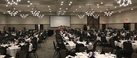 A large banquet hall with round tables, black chairs, and chandeliers. There is a stage with a screen at the front.