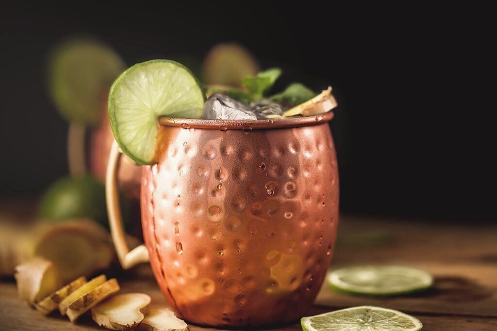 A copper mug with a lime slice and mint leaves, placed on a wooden surface with lime slices and ginger slices around it.