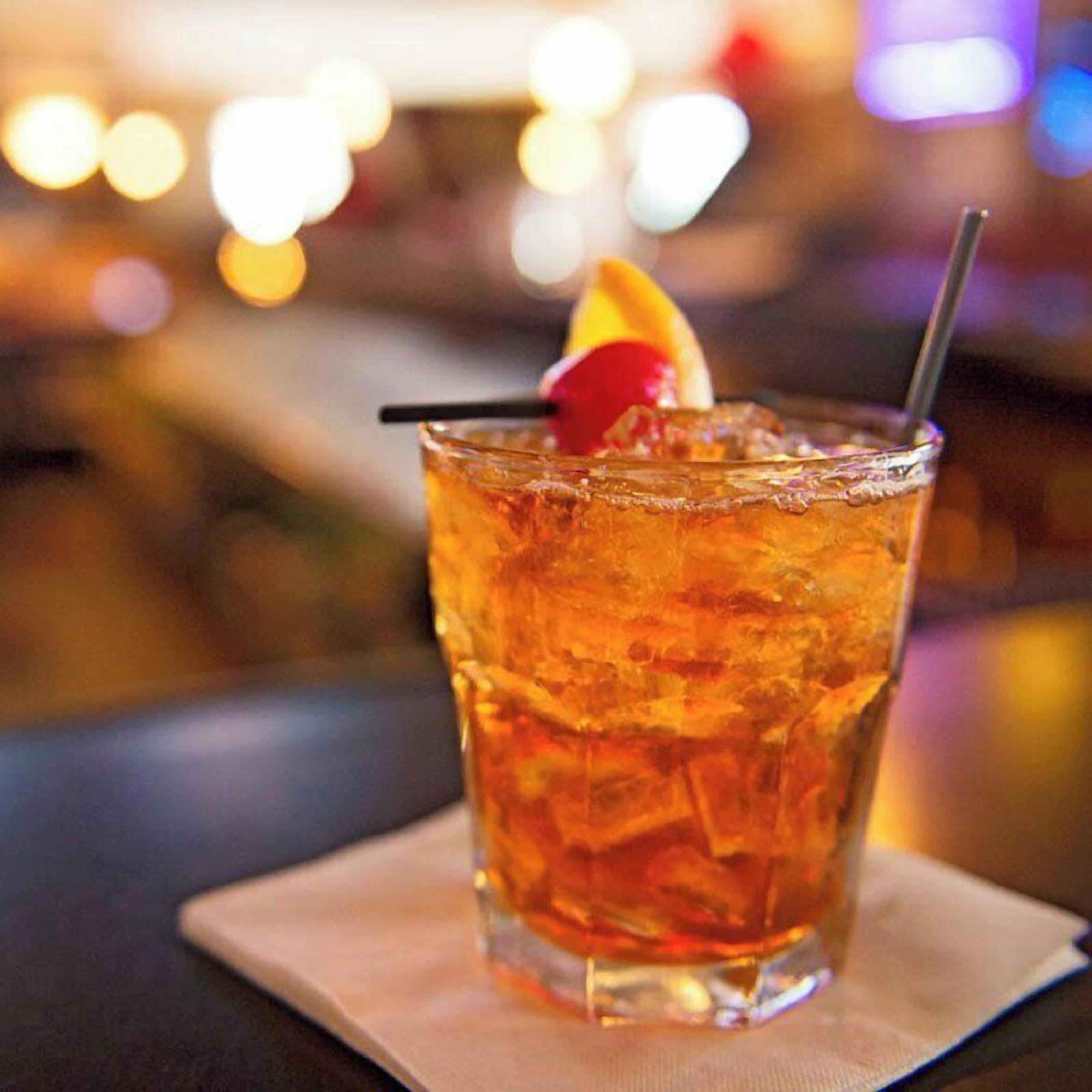 A glass of amber-colored beverage with ice cubes, a straw, and a garnish on a napkin.